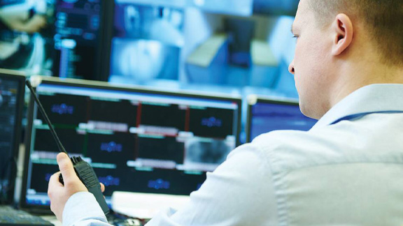 A security guard examining surveillance footage on a monitor while holding a walkie-talkie